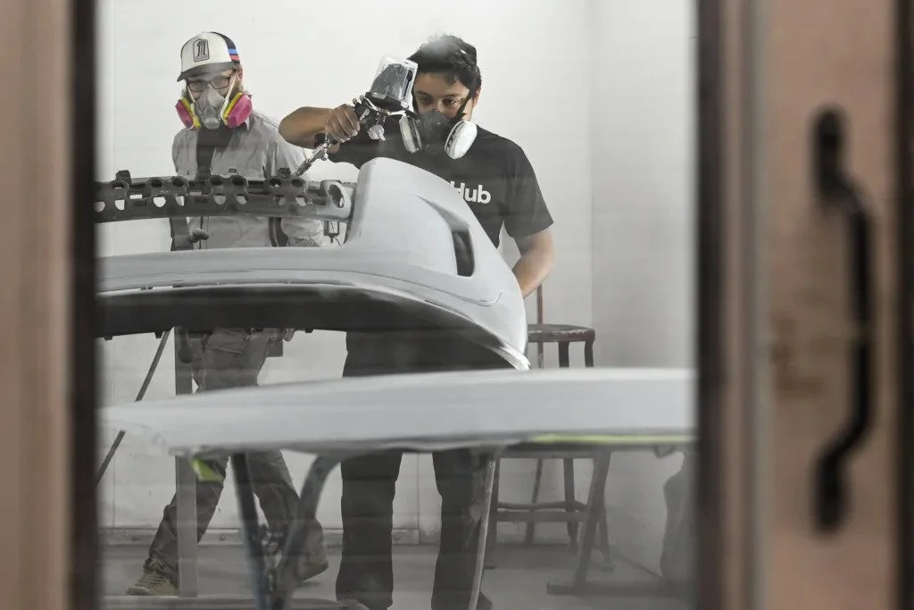 Two individuals working on a car part in a workshop, one is spray painting the car part while the other wearing a respirator mask observes.