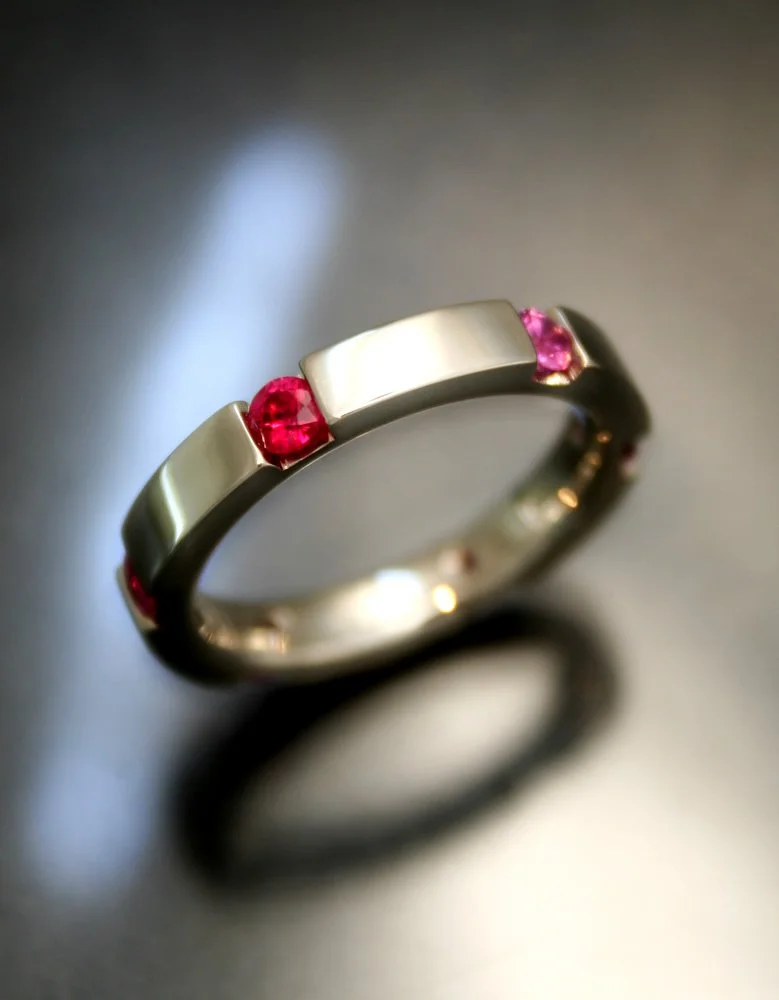 A silver ring with pink gemstones embedded on the top, resting on a reflective surface with soft lighting.