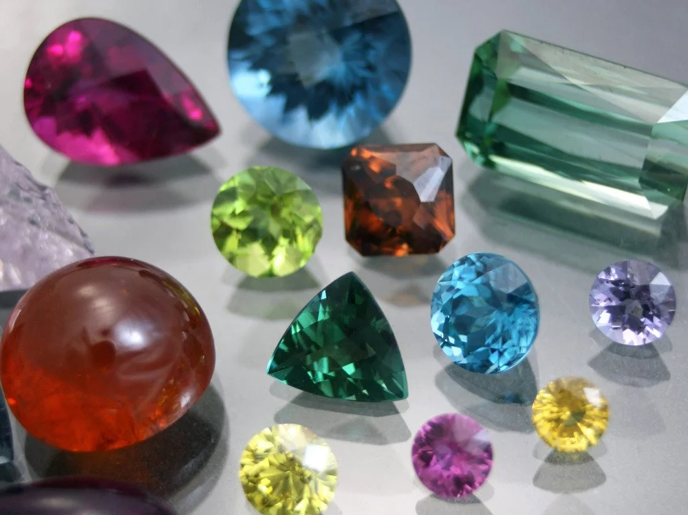 Assorted colorful cut gemstones on a gray surface, including round, oval, and rectangular shapes in pink, blue, green, yellow, purple, orange, and clear colors.