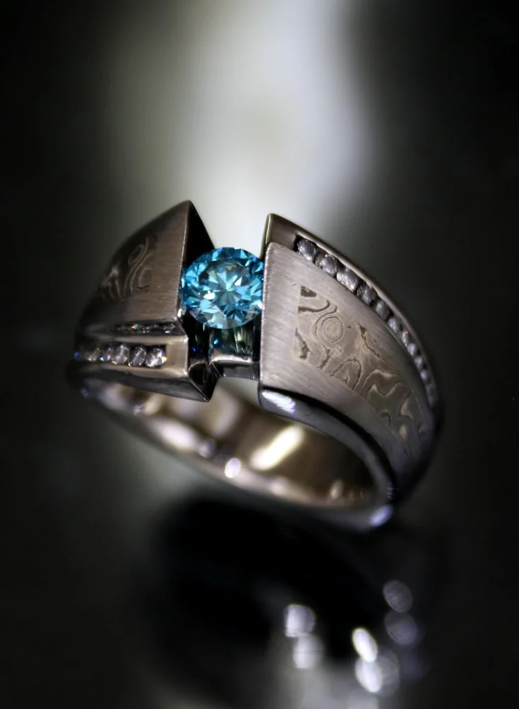 Close-up of a silver ring with a central blue gemstone and engraved designs, set against a dark background.
