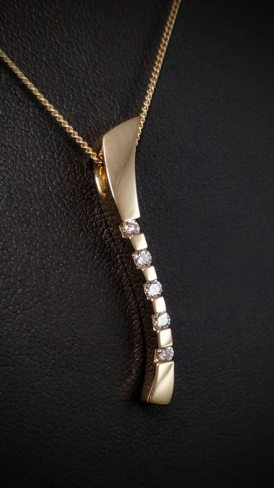 A gold necklace with a curved pendant and seven diamonds hanging from it, displayed against a black textured background.