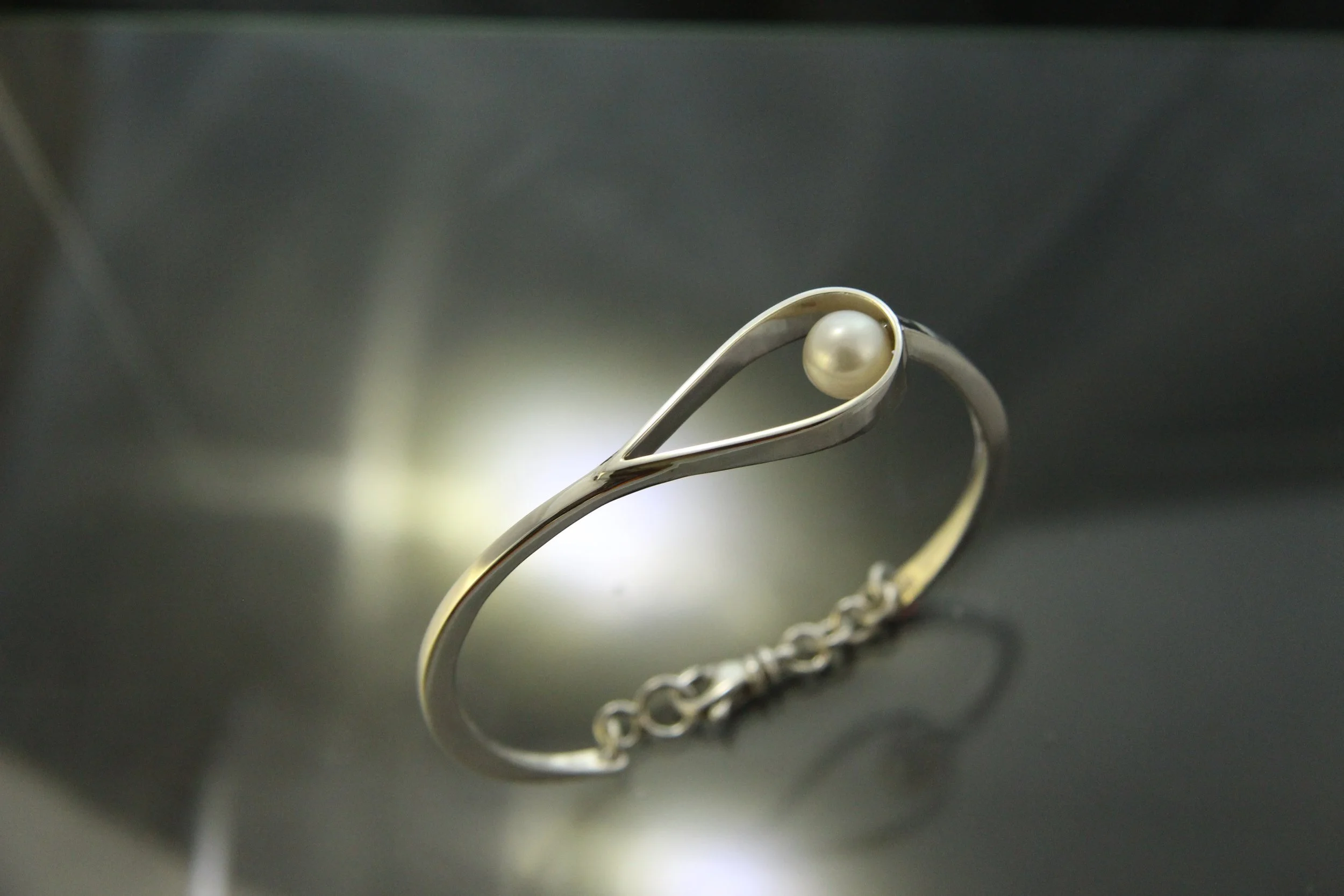 A silver bracelet with a pearl accent, placed on a reflective surface.