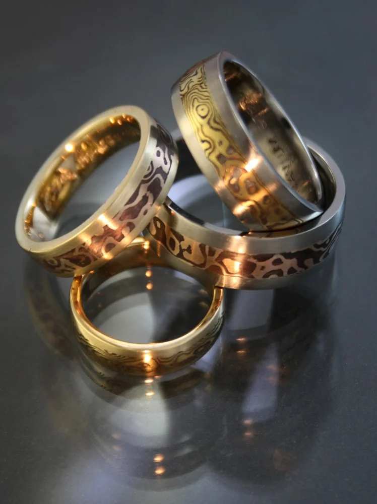 Four rings of different colors with engraved patterns, placed on a reflective dark surface.