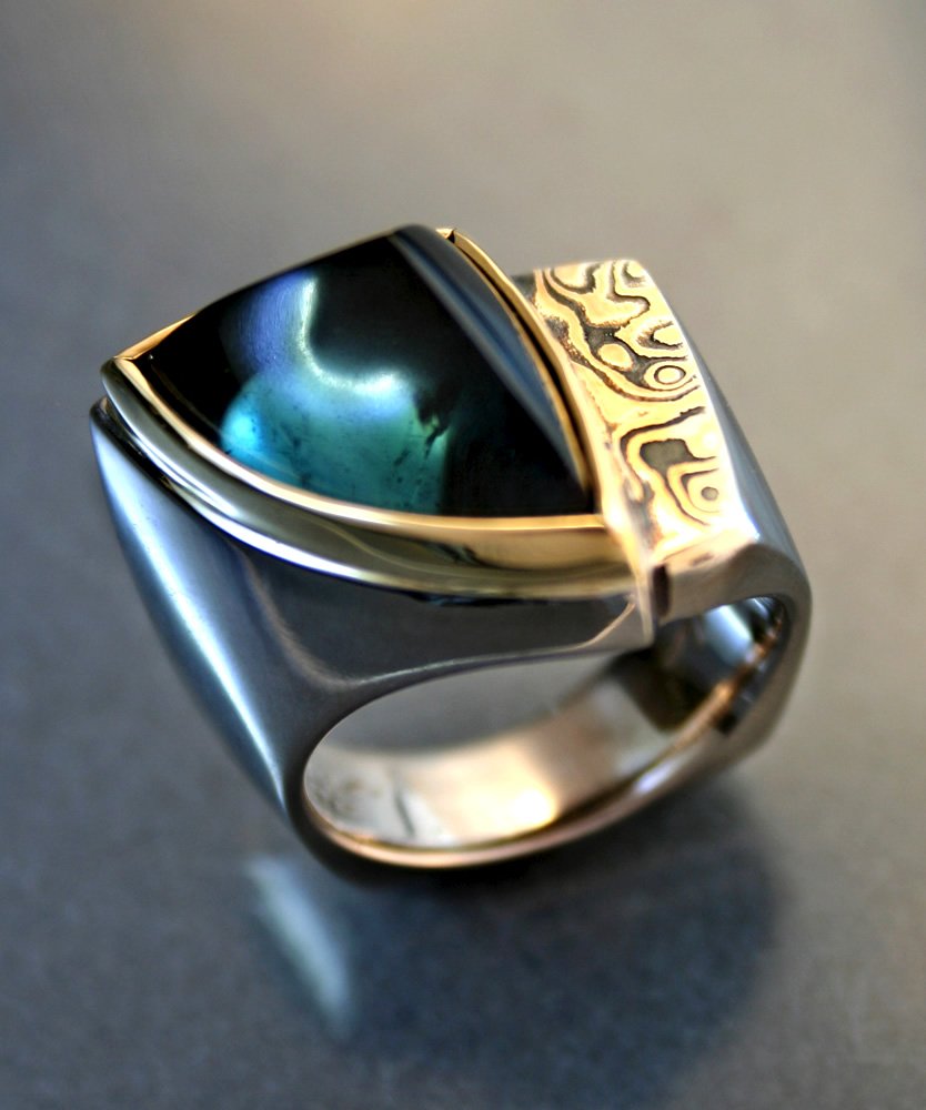 A silver ring with a large, rounded black stone set inside a gold bezel and an engraved gold band on top, resting on a dark surface.