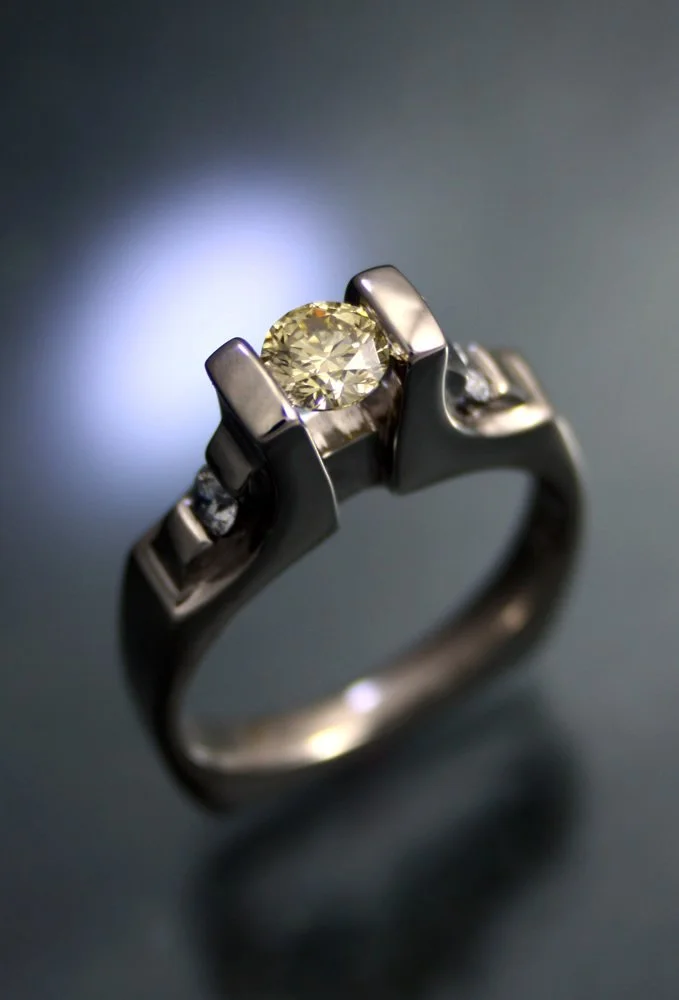 A close-up of a silver ring with a large central yellow gemstone and two smaller clear stones on each side, on a dark background.