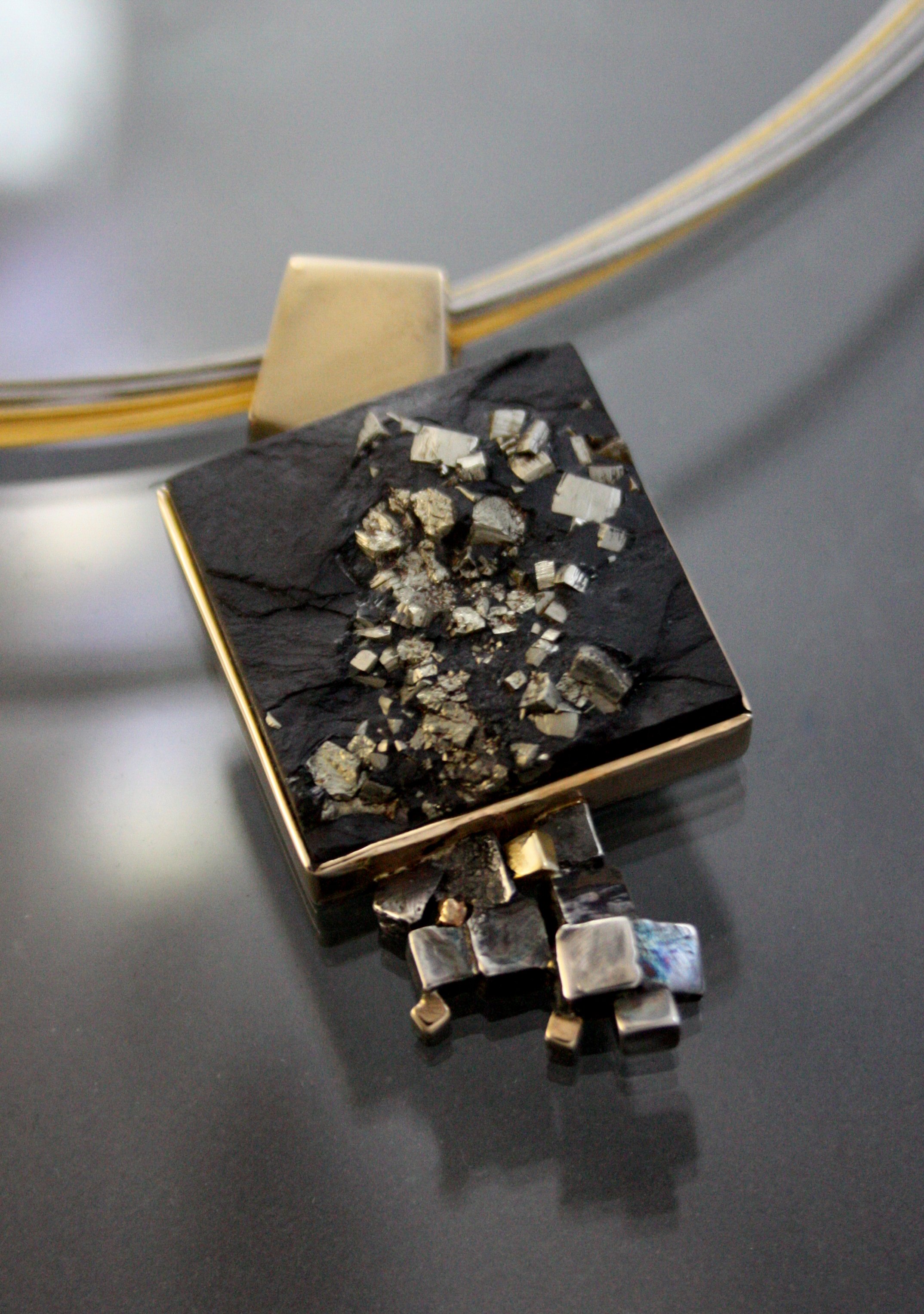 Close-up of a jewelry piece with a black and gold geometric design, featuring silver-colored metallic stones and a chain with square and rectangular links.