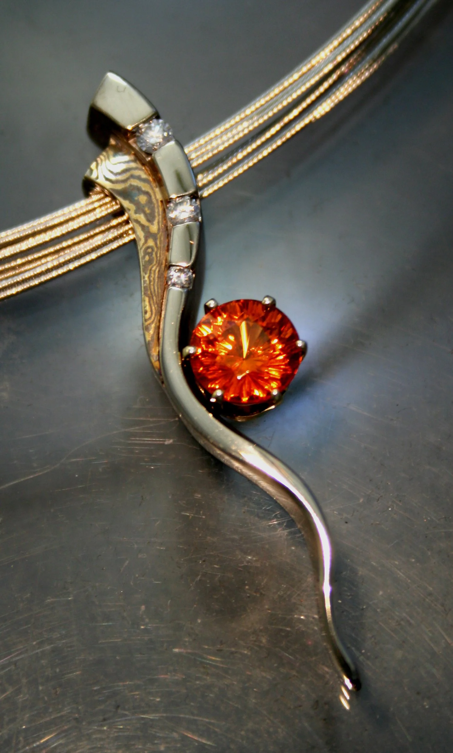 Close-up of a silver and gold necklace with a large orange gemstone and small diamonds on a metallic surface.
