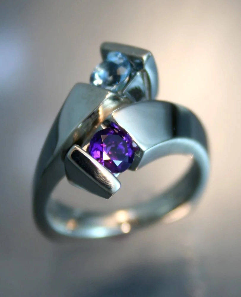 A silver ring with two stones, one light blue and one purple, set in a modern, sculptural design.
