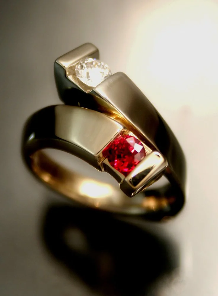 Close-up of two metallic rings with embedded gemstones, one with a clear diamond and the other with a red gem, on a reflective surface.