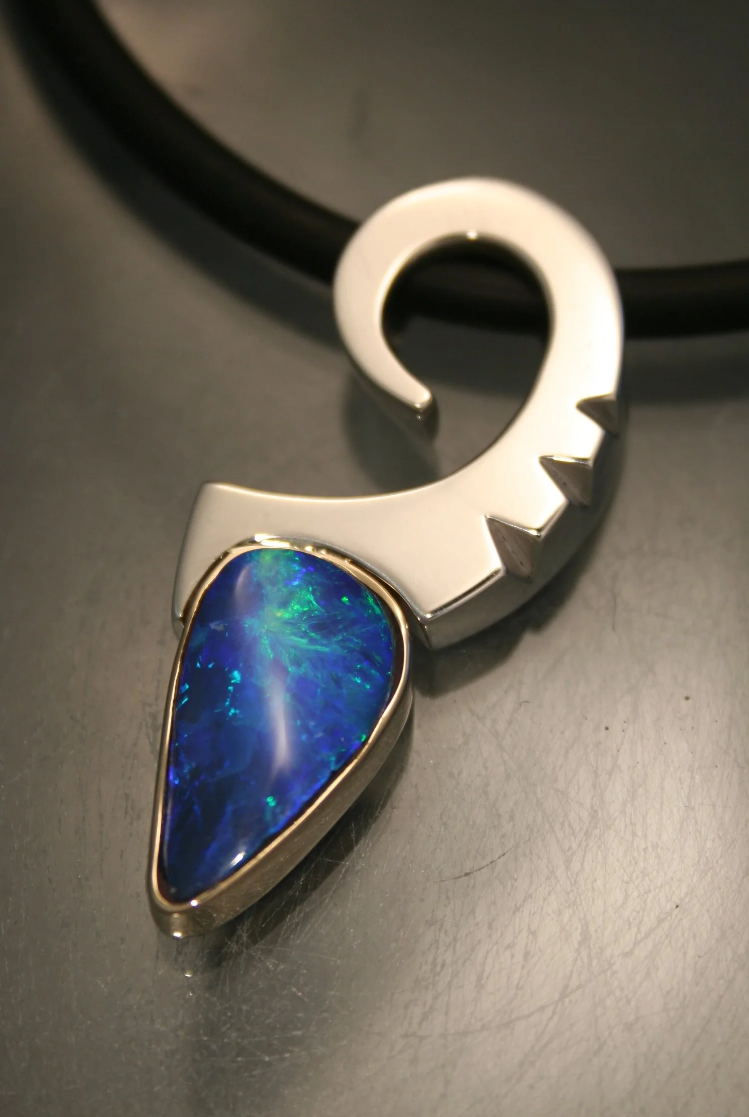 A silver jewelry piece with a large, teardrop-shaped blue opal gemstone set in it, lying on a dark surface with part of a black circular object in the background.