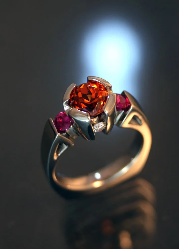 A silver ring featuring a large orange gemstone flanked by two smaller pink gemstones, with a small clear stone beneath the orange gemstone, against a dark background with reflective surface.