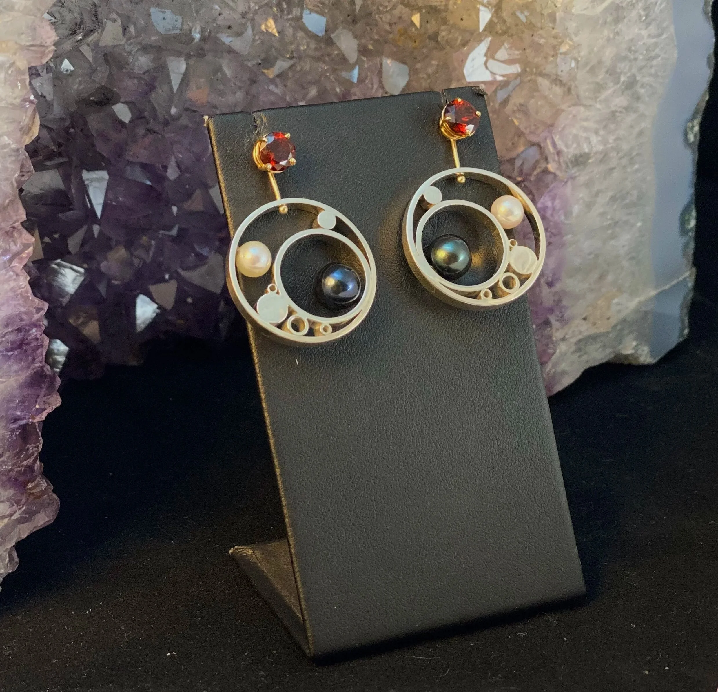 Pair of earrings with circular design, black and white pearls, and red gemstone accents, displayed on a black stand with purple mineral crystals in the background.