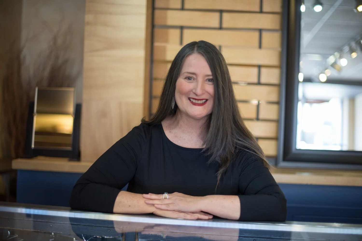 A woman with dark brown hair and red lipstick is smiling and sitting at a table in a modern, well-lit interior with wooden paneling.