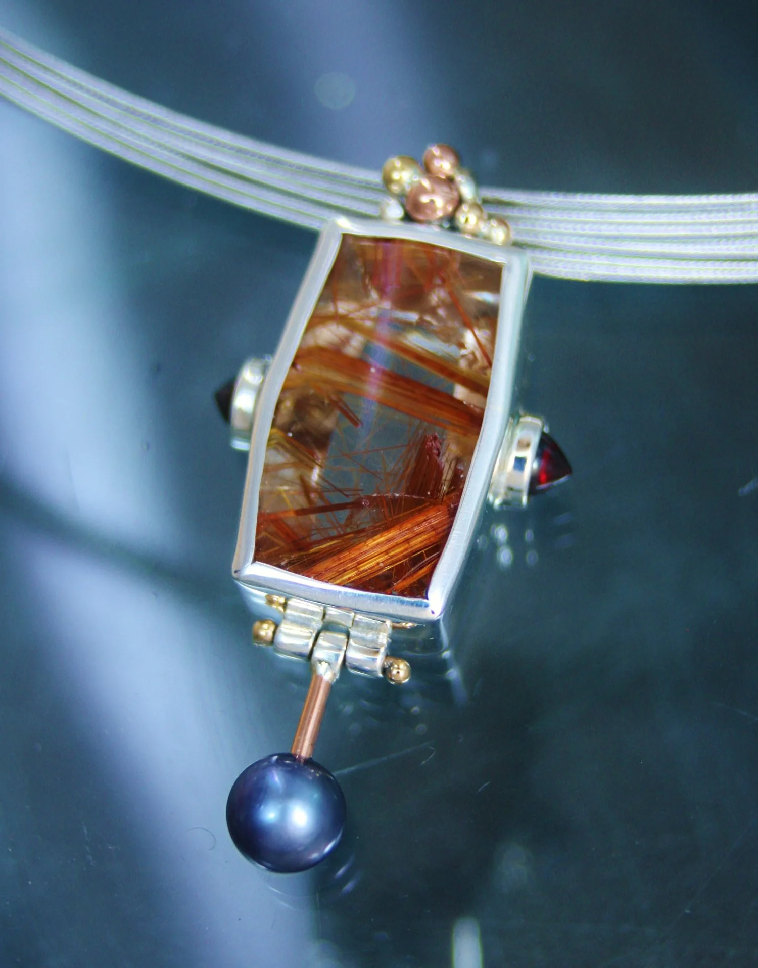 A close-up of a jewelry piece with a rectangular amber-colored stone and metallic accents, attached to a layered silver necklace with spherical beads.
