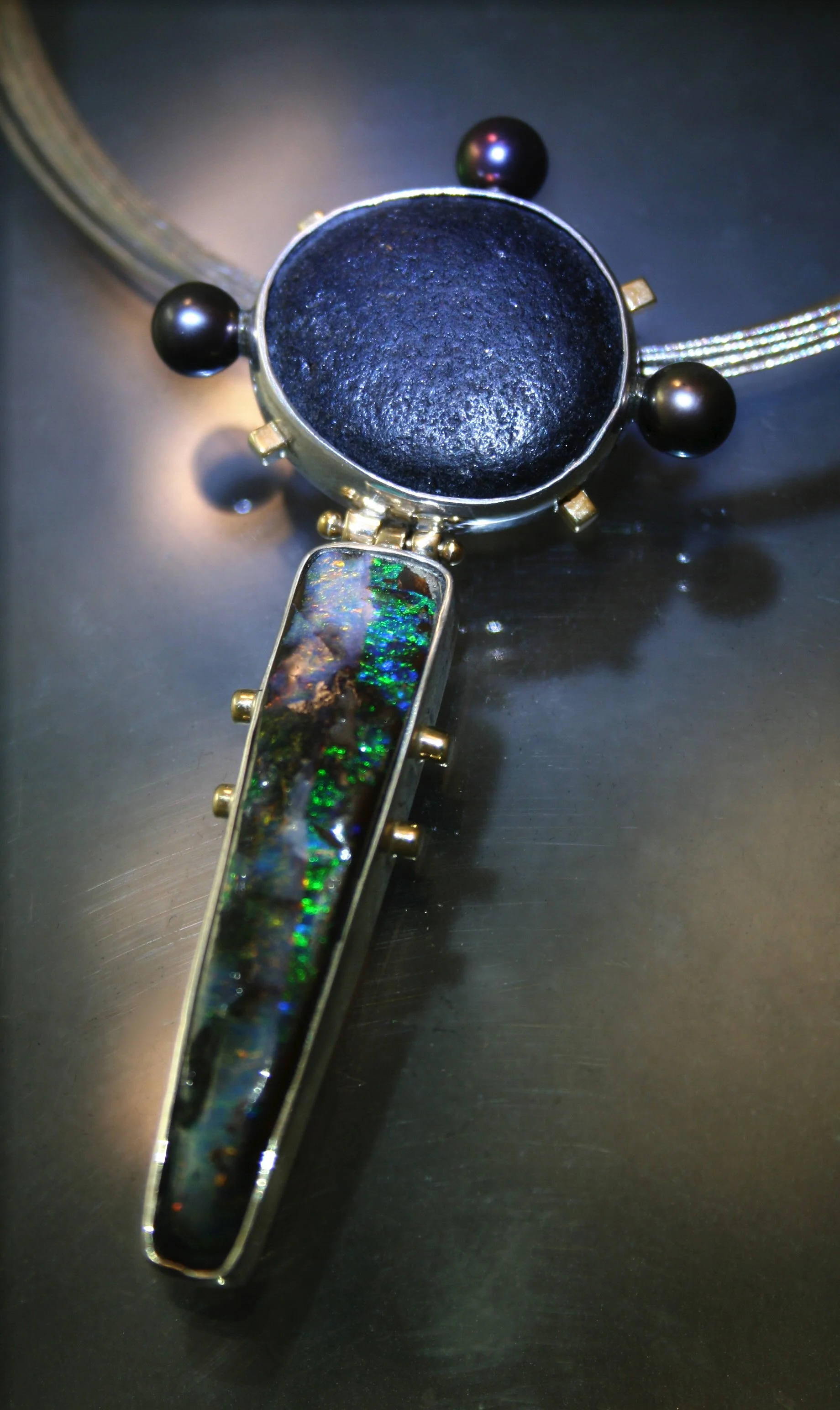 A pendant necklace with a large, round, black gemstone and a long, narrow, rectangular pendant featuring an iridescent, multicolored inlay, set in a metal frame with small decorative metal elements.