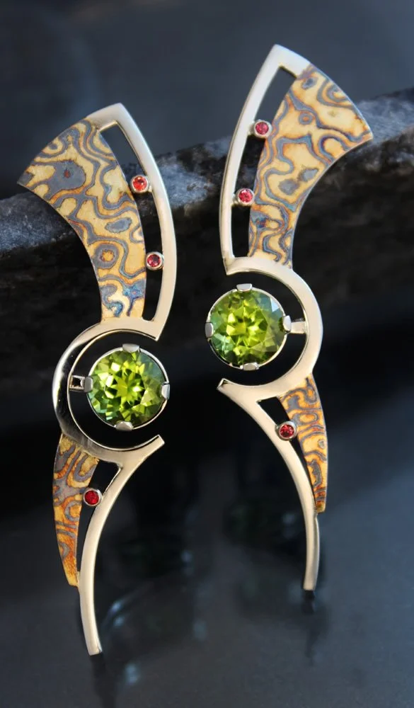 A pair of modern earrings with a unique geometric design, featuring oval-shaped green gemstones and red accent stones set in silver and gold-toned metal with intricate patterned details.
