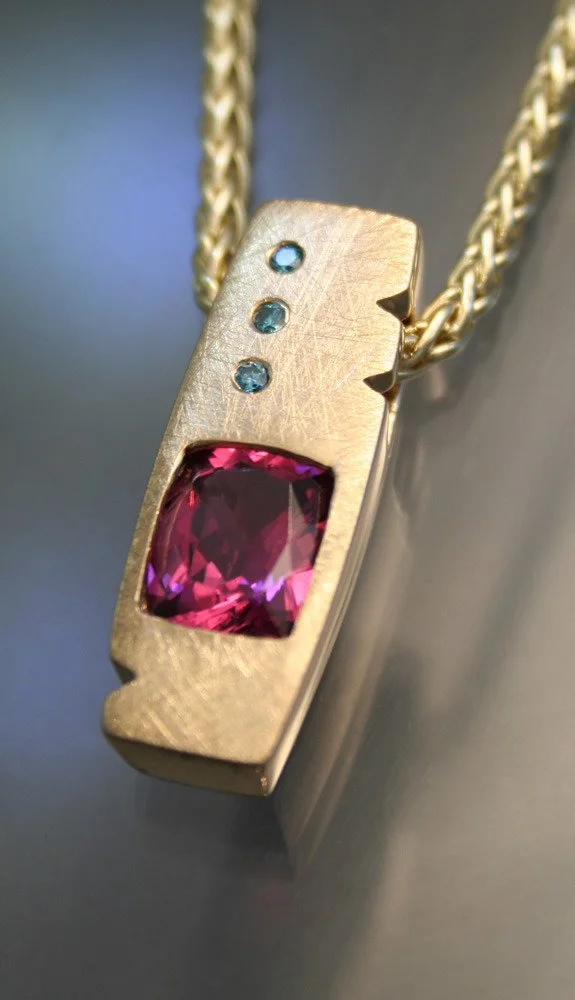 A gold pendant necklace with three small blue gemstones and one large pink gemstone on a chain.