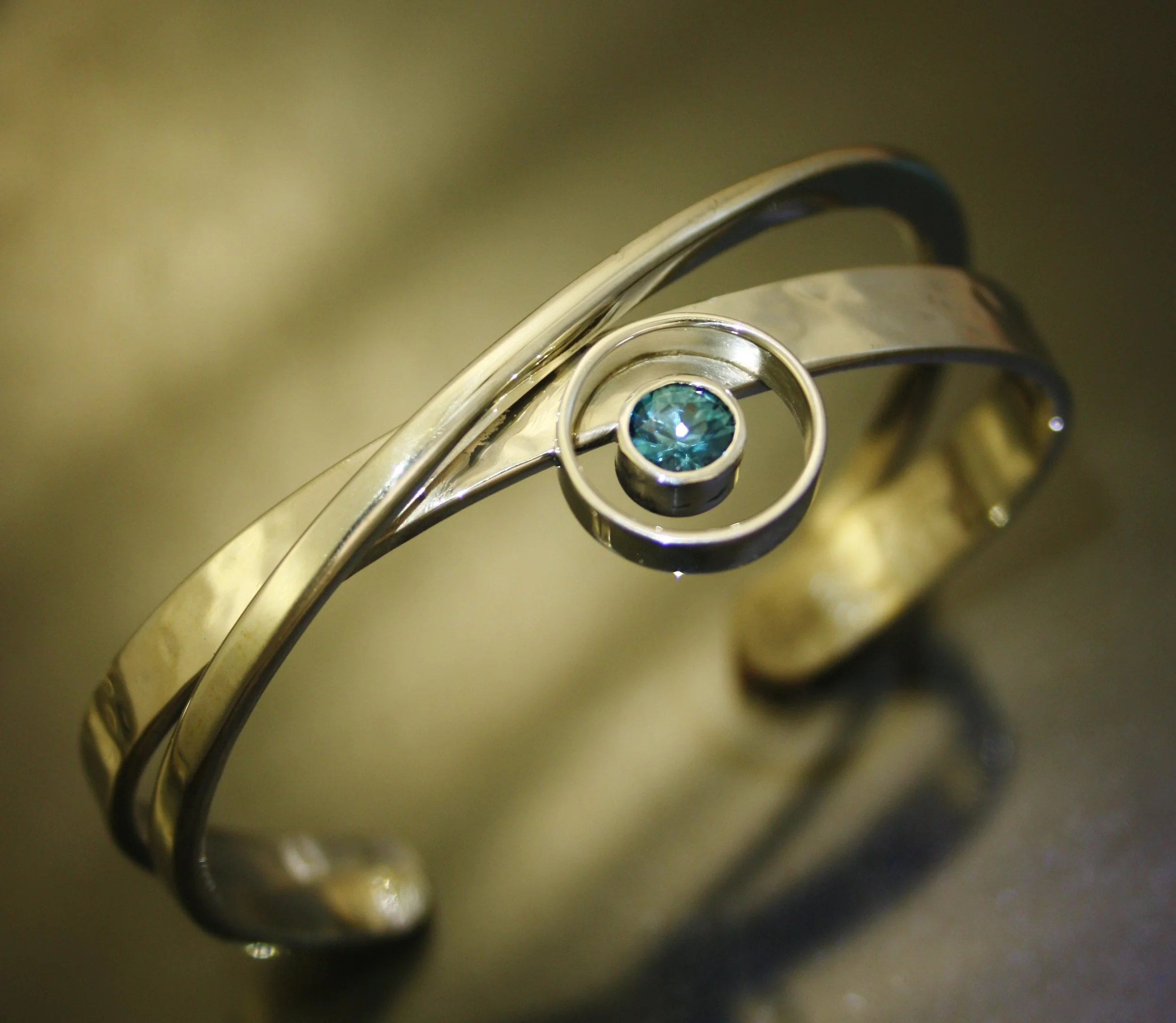 Close-up of a silver ring with a spiral design and a blue gemstone at the center, on a reflective surface.