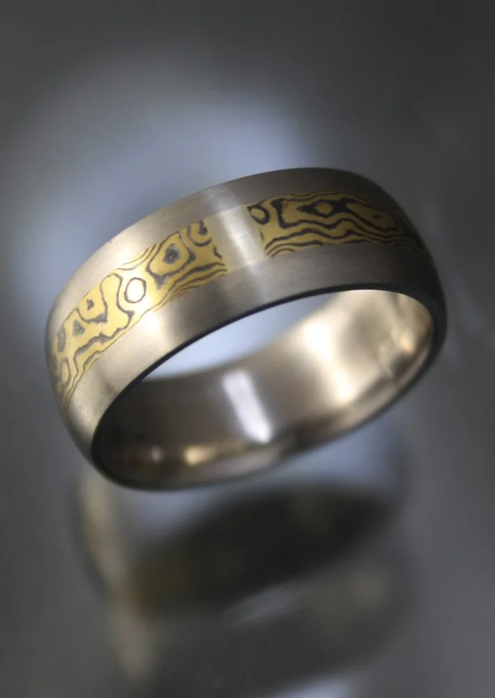 Close-up of a silver rings with gold patterned inlay, resting on a reflective dark surface.
