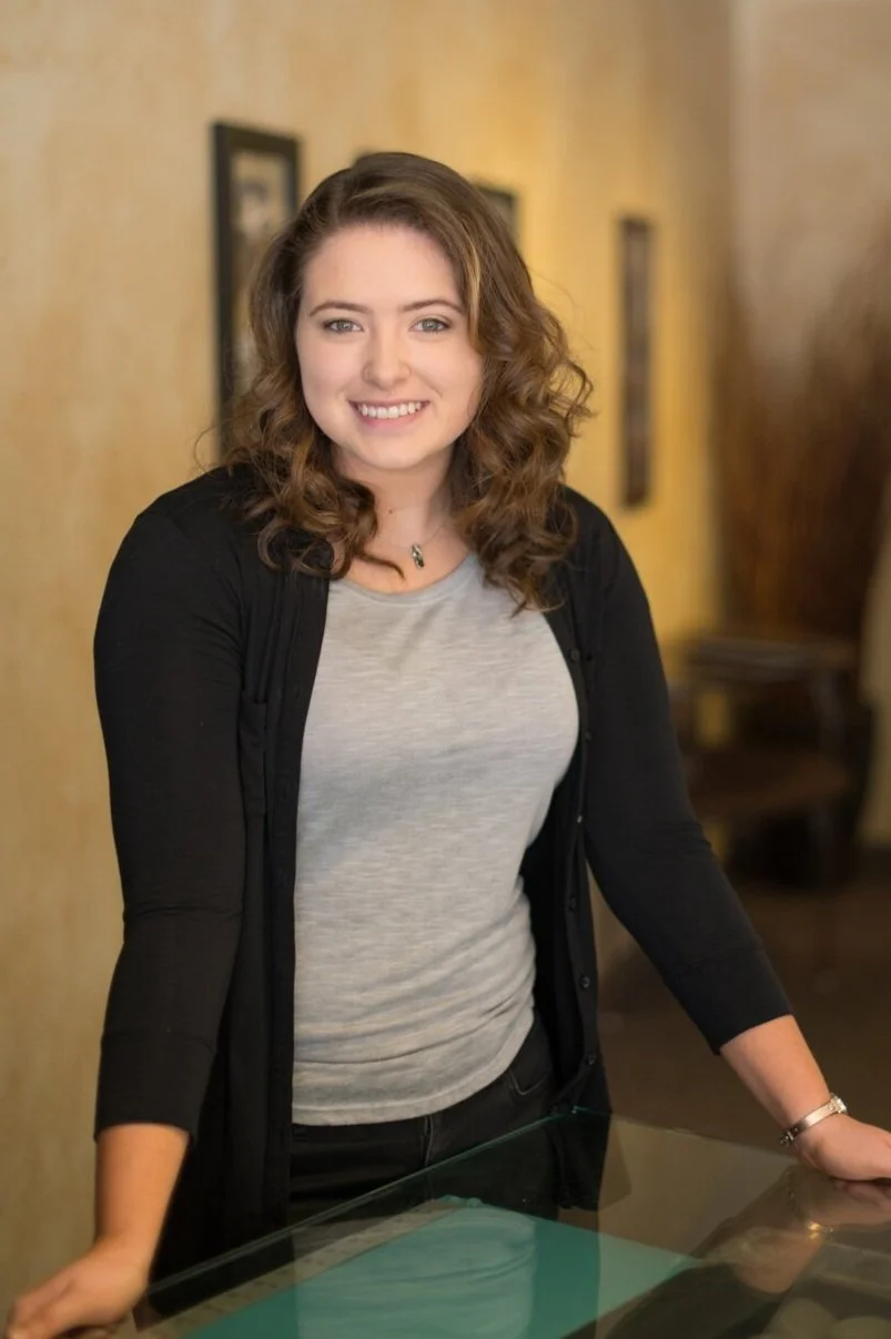 A young woman with shoulder-length curly brown hair, smiling, standing indoors near a glass table with a yellow wall and framed pictures in the background.