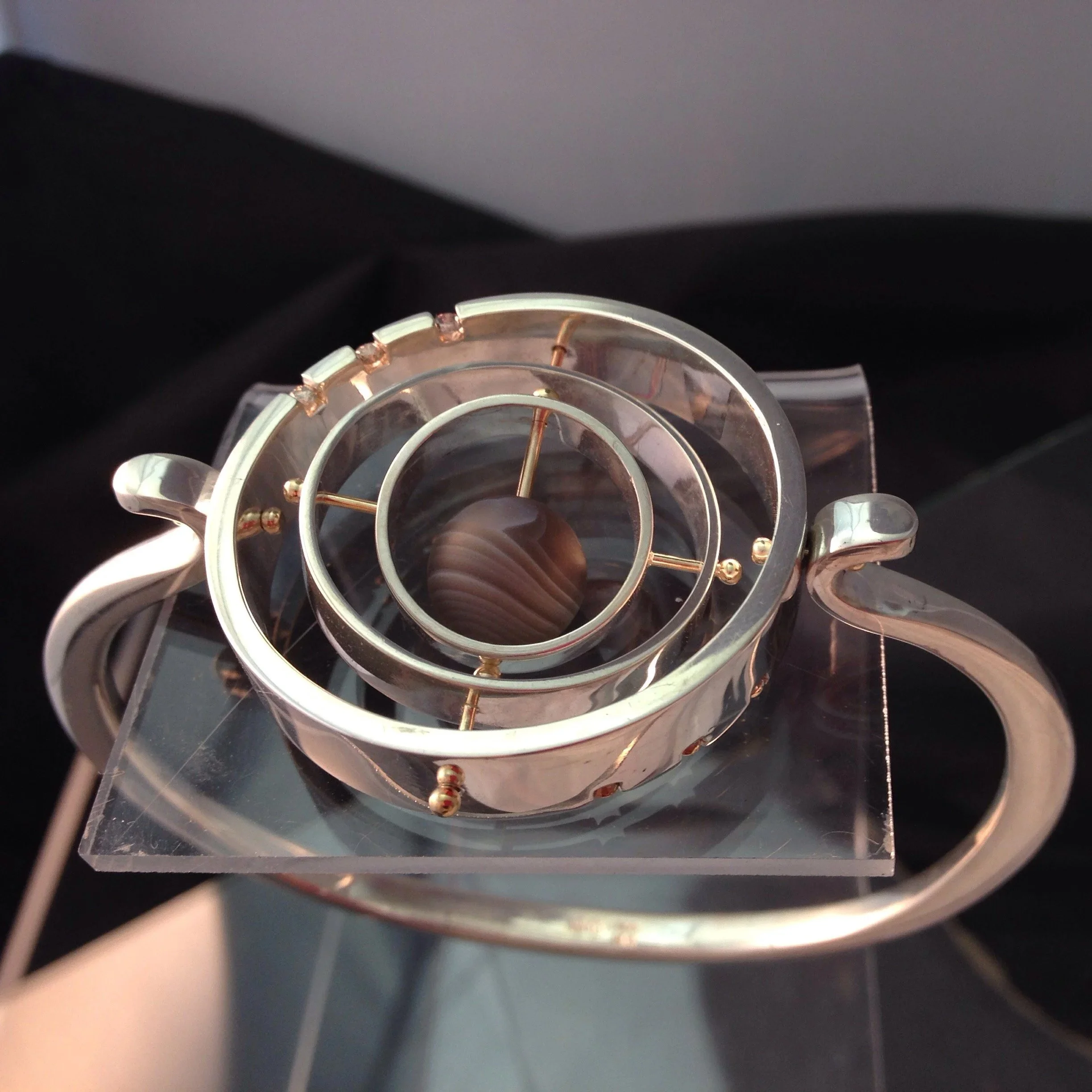 A metallic spiral-shaped sculpture with circular rings, placed on a transparent stand, reflecting a marble stone inside a clear glass case.