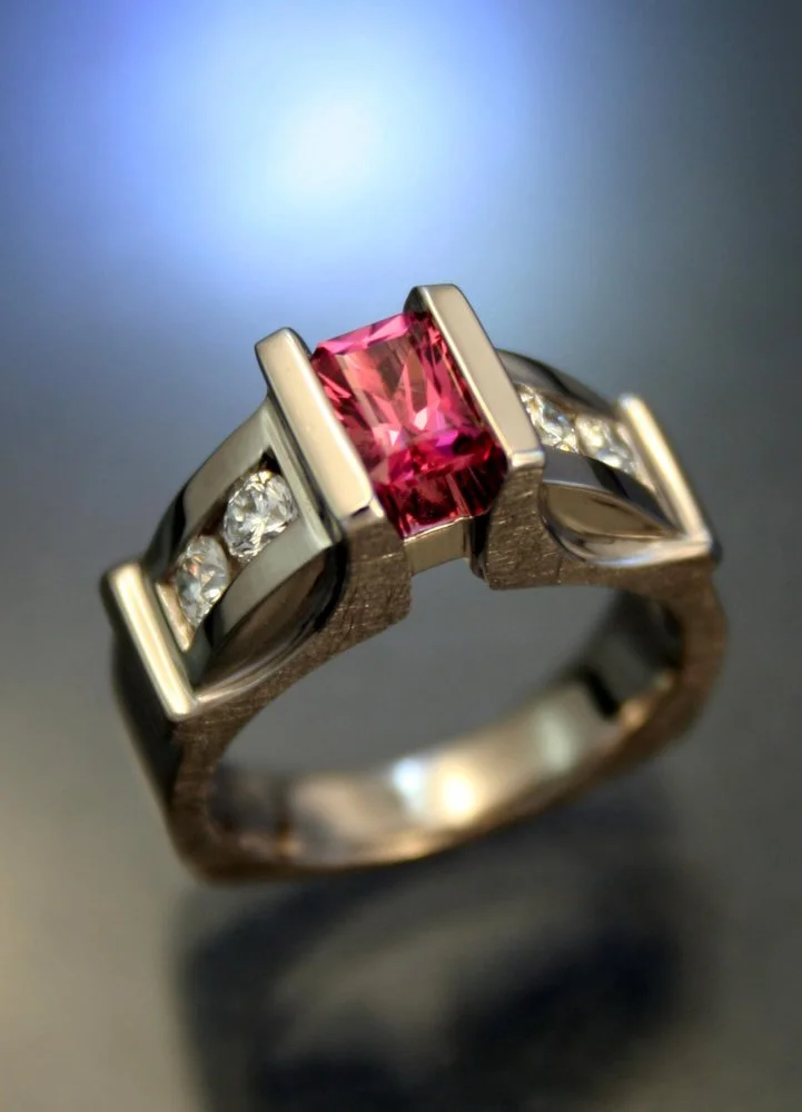 A close-up of a silver ring with a rectangular pink gemstone and small diamonds on either side, set against a dark background with a blurred light in the upper left corner.