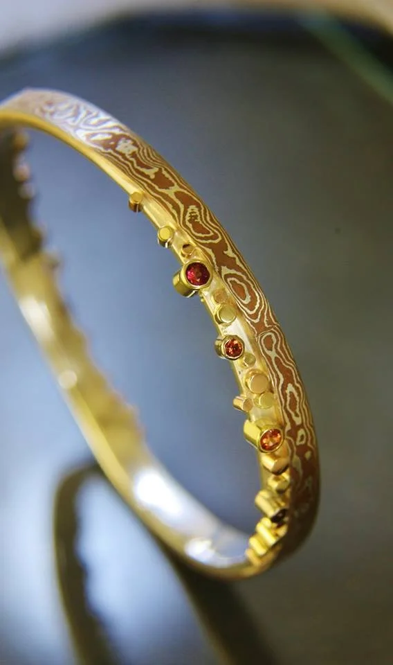 Close-up of a gold bracelet with a textured pattern and small embedded red gemstones, resting on a dark reflective surface.
