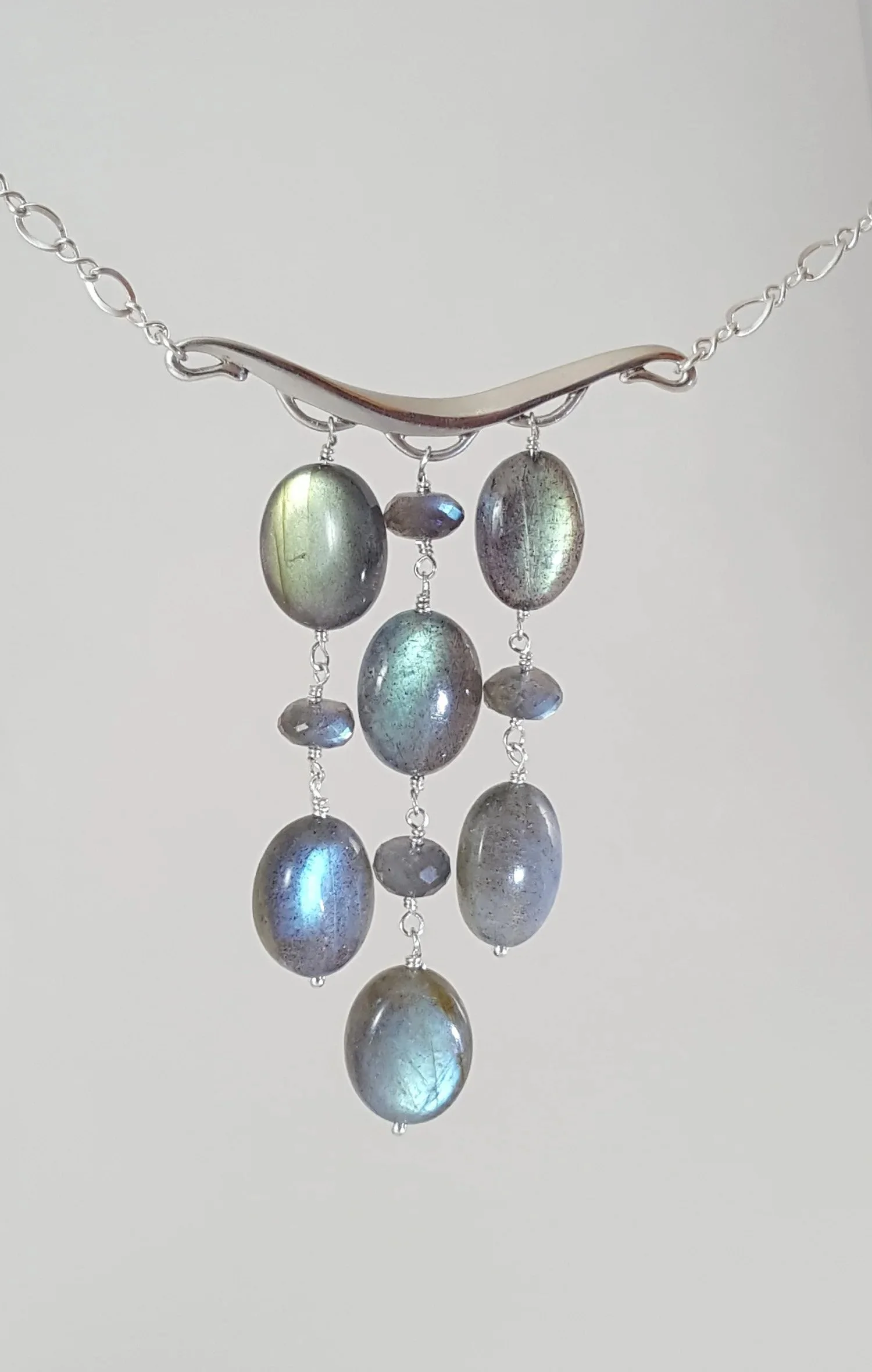 Silver necklace with a curved bar and hanging oval and round metallic beads, some with iridescent reflections.