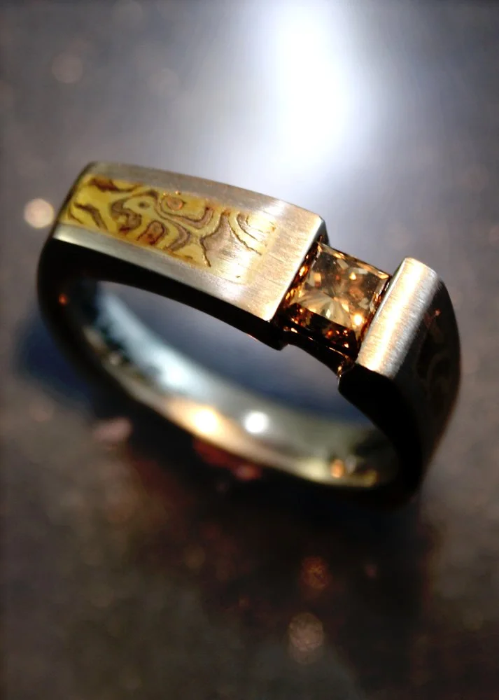 Close-up of a modern men's ring with a brushed metal finish, featuring a small gemstone and an intricate woodgrain pattern on the top inlaid section.