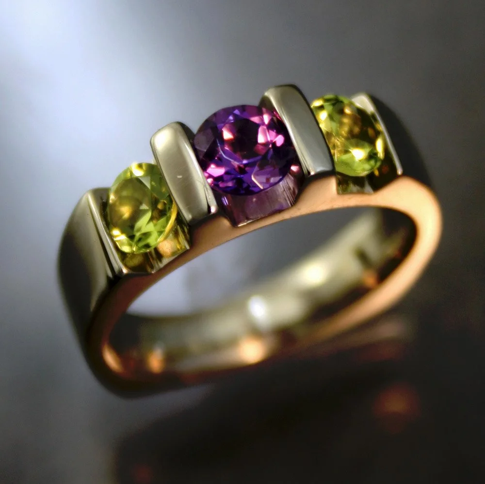 Close-up of a ring with a purple gemstone in the center flanked by two yellow-green gemstones, set in metal band.