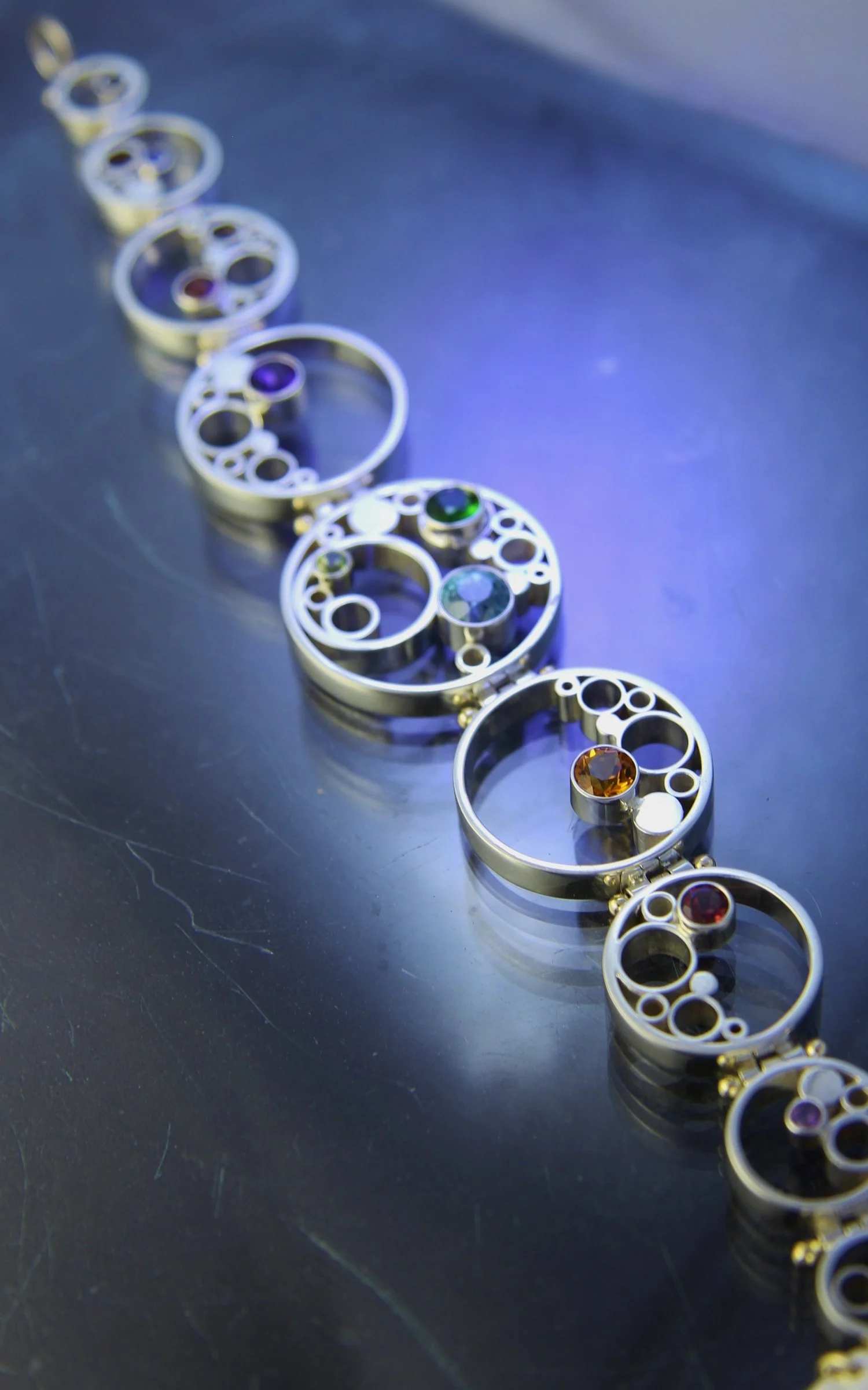Close-up of interconnected metallic rings with colorful gemstones inside, arranged in a diagonal line on a reflective dark surface.