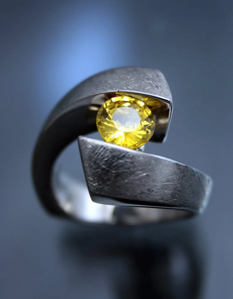 Close-up of a silver ring with a yellow gemstone set in a unique, twisted design on a dark reflective surface with a blurred dark background.