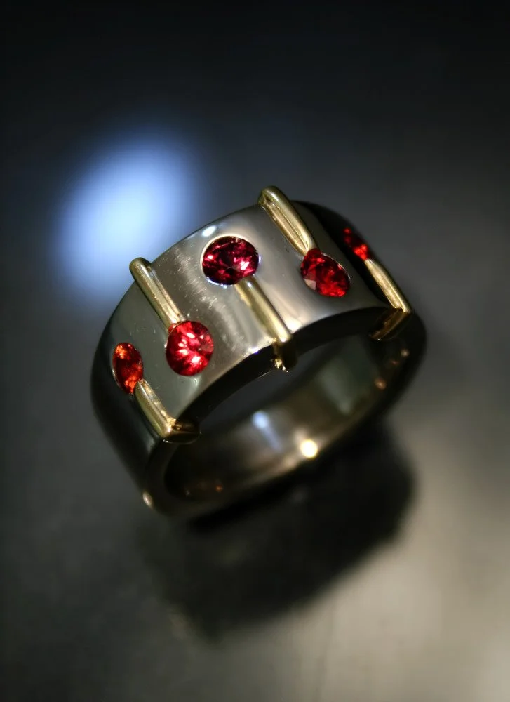 A silver ring with gold accents and five red gemstones, resting on a dark surface under focused lighting.
