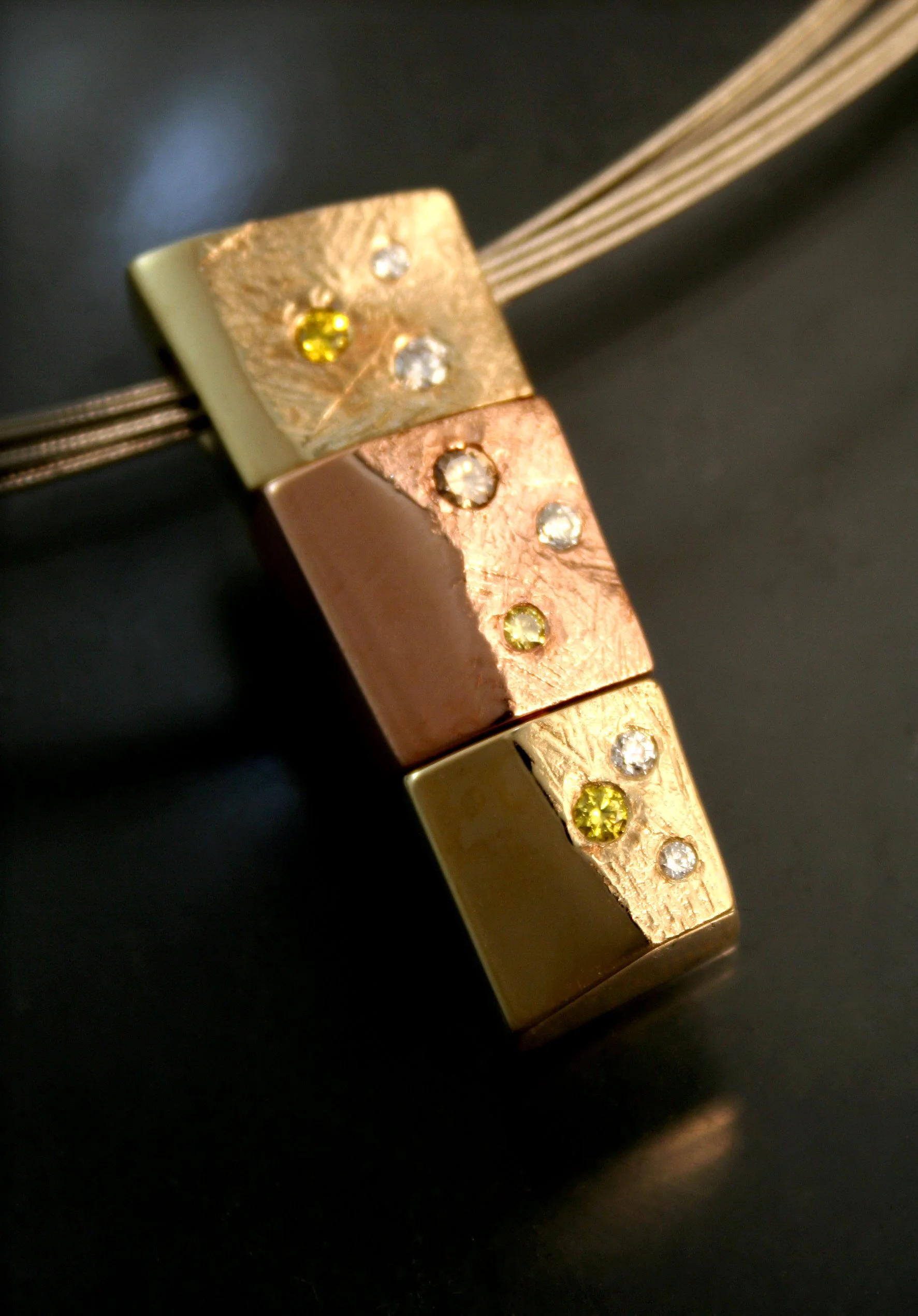 Three metallic cubes, one cream-colored, one rose gold, and one brass, each with embedded rhinestones, stacked on a black background, with strings attached to the cream-colored cube.