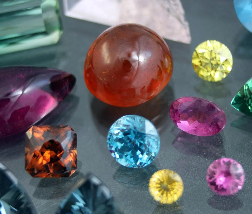 Various colorful gemstones and crystals on a black surface, including a large orange sphere, a purple teardrop, a blue round, a pink oval, a yellow round, and a square-shaped orange gem.
