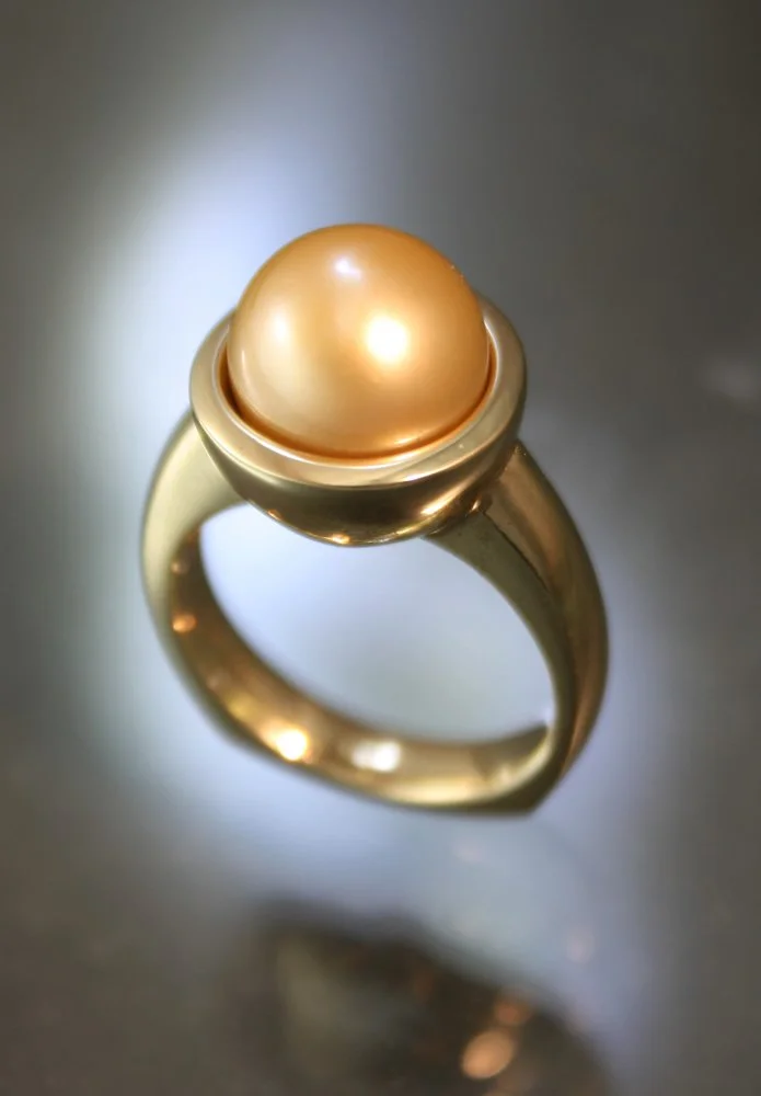 Gold ring with a large golden pearl set in a gold bezel, photographed from above on a gray background.