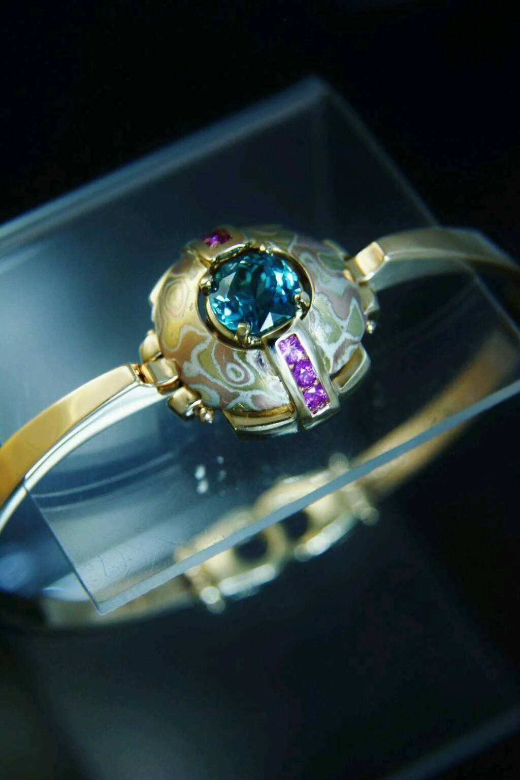 Close-up of a gold jewelry piece with a large blue gemstone and pink gemstones, displayed on a transparent stand.