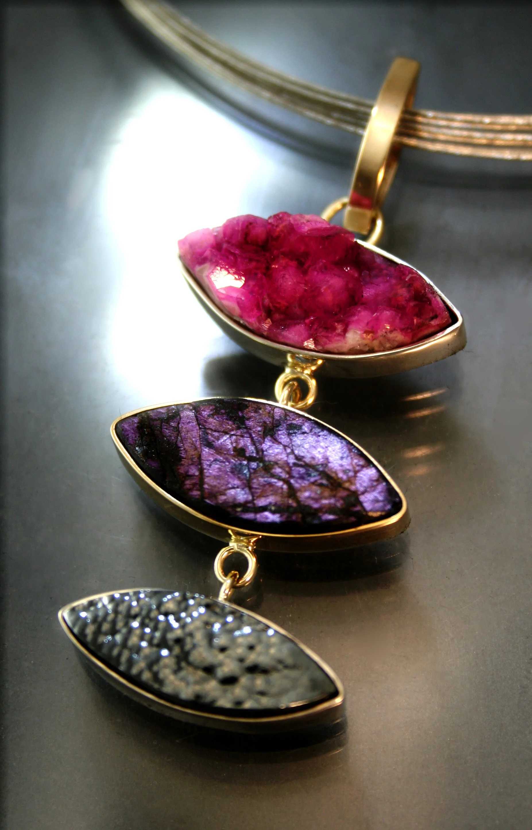 A jewelry piece with three large, irregularly shaped stones set in gold, each stone a different color: pink, purple, and black, arranged in a vertical line.