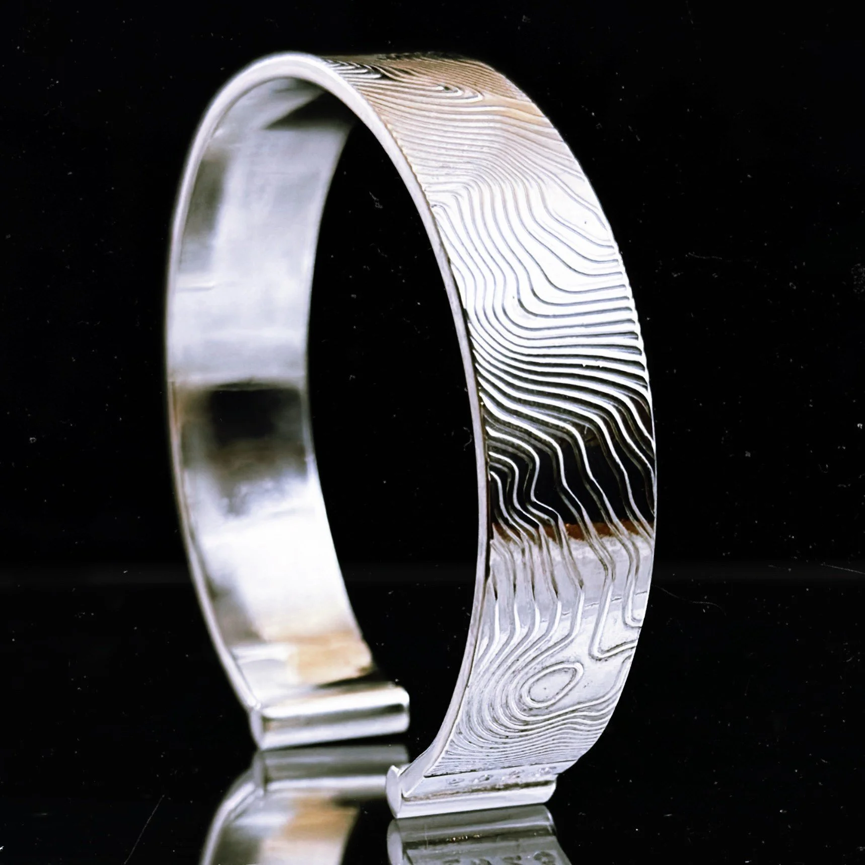 Close-up of a silver ring with engraved patterns, reflecting on a black surface.