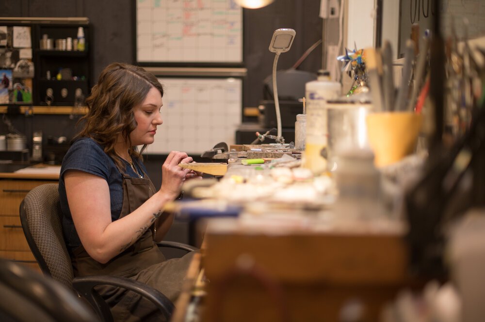 A woman working on jewelry at a cluttered workbench in a workshop