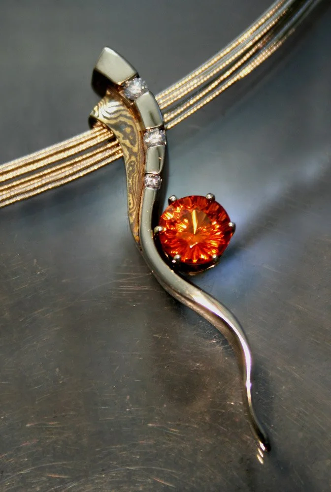Close-up of a silver and gold pendant necklace featuring a curved design with a large orange gemstone and small diamonds, resting on a metallic surface.