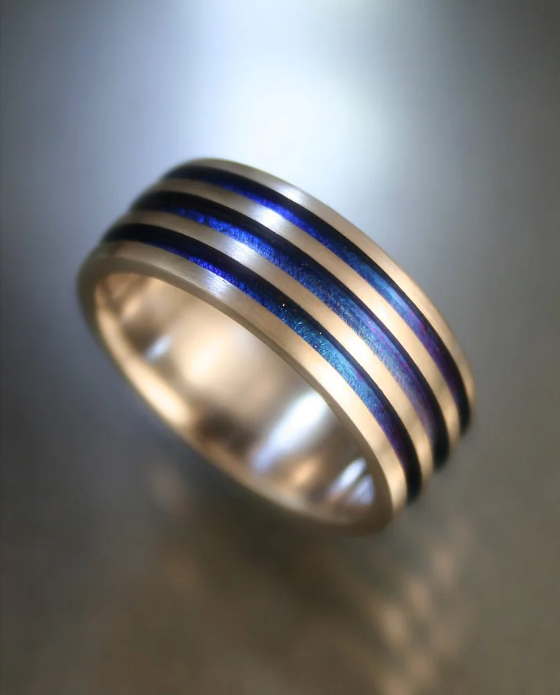 Close-up of a gold ring with four parallel black and blue stripes, resting on a reflective surface.