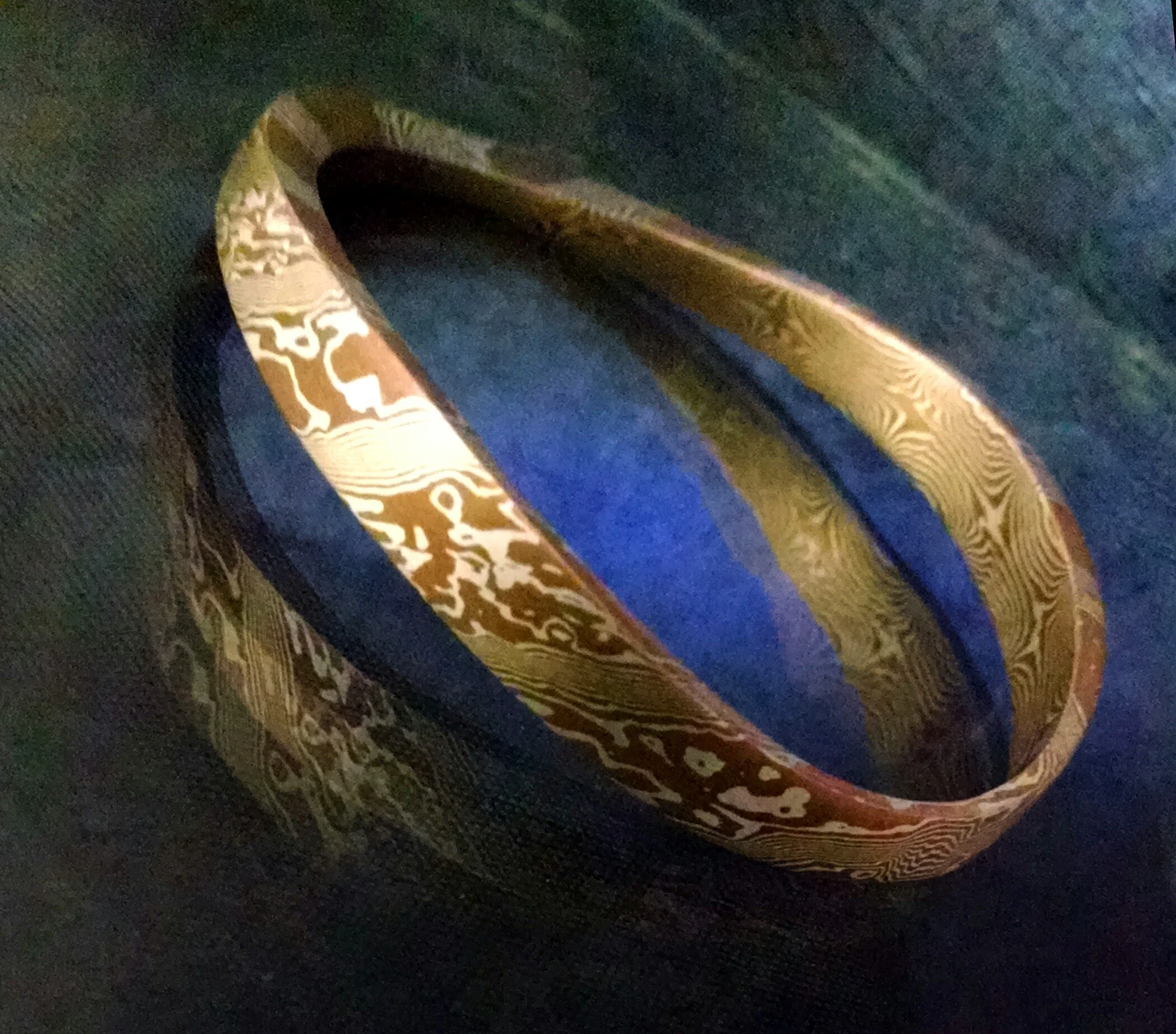 A close-up view of a metallic bracelet resting on a dark surface. The bracelet has intricate gold-colored swirling patterns on its surface.
