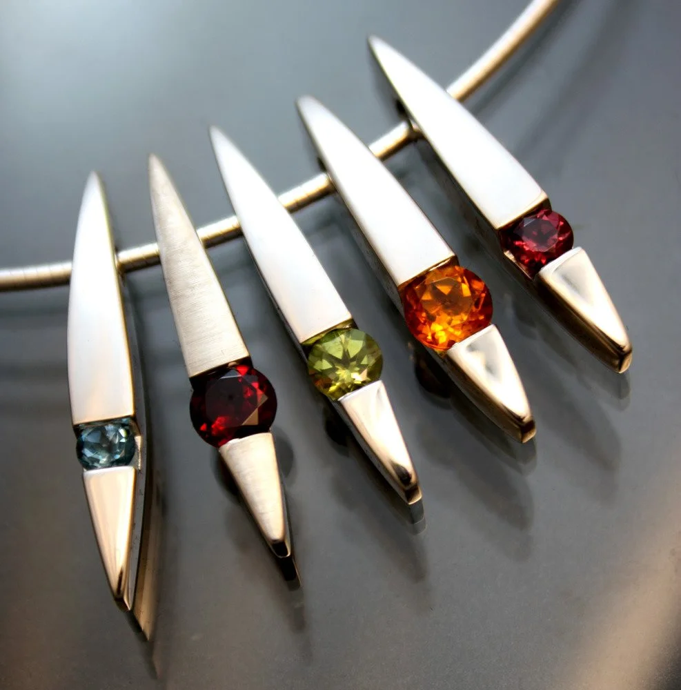 A silver necklace with five silver pendants, each featuring a colorful round gemstone in the center, with colors ranging from blue, red, green, orange, to red.