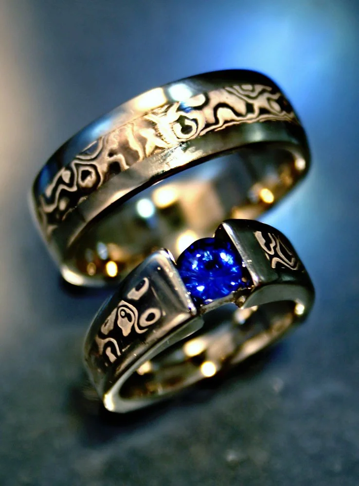 Close-up of two silver rings with intricate black patterns; one ring features a blue gemstone. Both rings have unique, engraved designs and are displayed on a dark background.