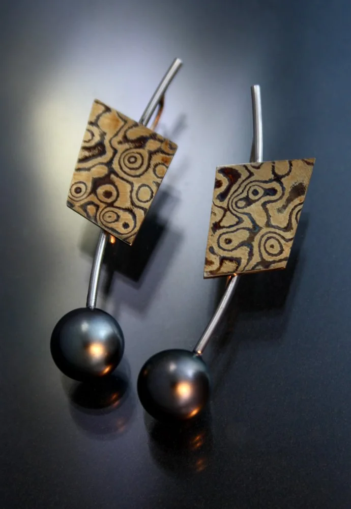 Black earrings with rectangular patterned gold and black designs and large black beads at the bottom, placed on a dark surface.