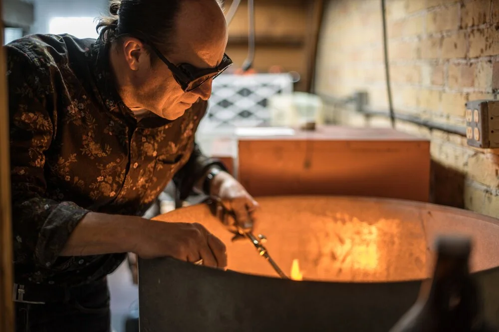A man wearing glasses and a floral shirt is heating a metal object in a large, industrial furnace.