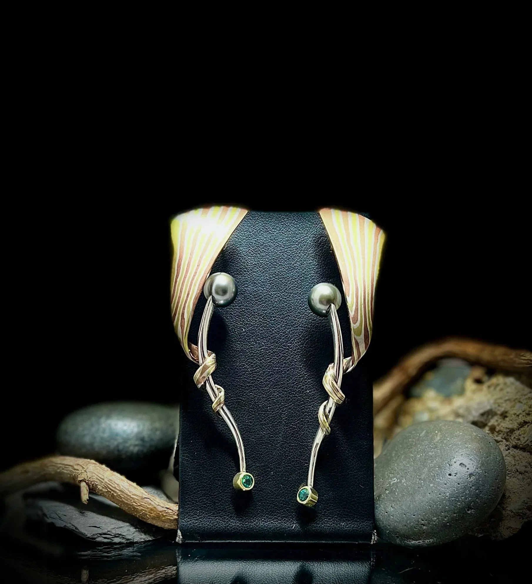 Silver earrings with green gemstones hanging from twisted silver wires, displayed on a black jewelry stand with a colorful scarf around the top, surrounded by smooth stones and twigs.