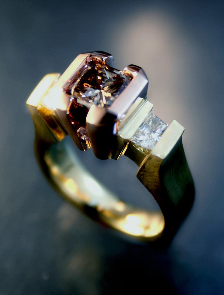 A gold ring with a large square-cut diamond at its center, accented by smaller diamonds on the band, photographed with a dark blurred background.