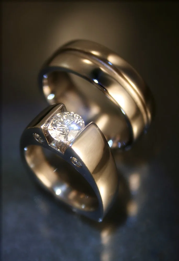 Close-up of two silver rings, one with a prominent diamond, on a dark reflective surface.